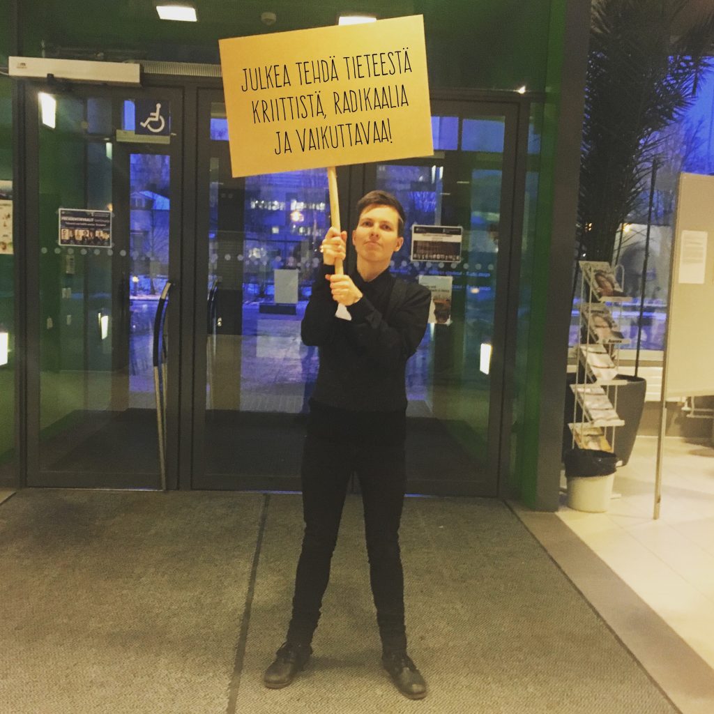 Henkilö seisoo kyltti kädessään. Kyltissä lukee: "Julkea tehdä tieteestä krittistä, radikaalia ja vaikuttavaa".