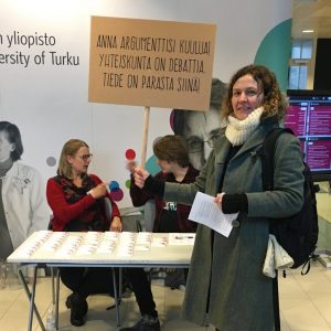 Henkilö seisoo kyltti kädessään. Kyltissä lukee: "Anna argumenttisi kuulua! Yhteiskunta on debattia, tiede on parasta siinä."