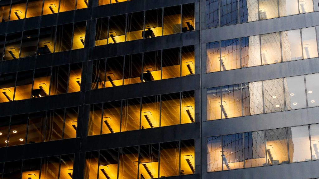 Abstrakti kuva modernin rakennuksen ulkoseinästä, jossa lasiseinien takana näkyy rakennuksen sisällä päällä olevat valot.
