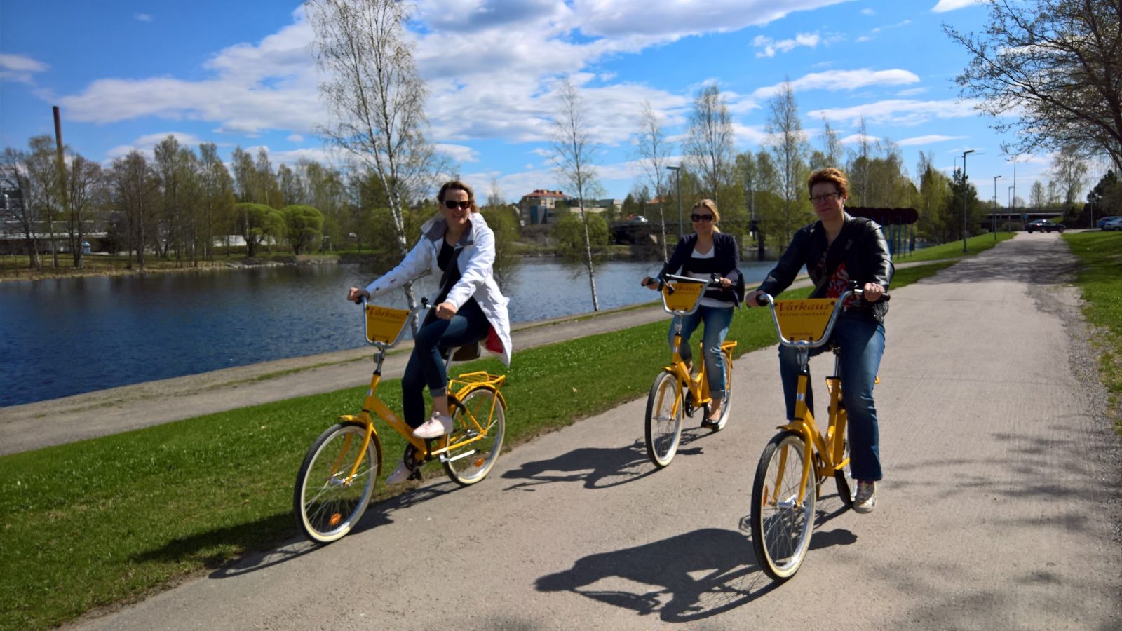 Kolme henkilöä pyöräilevät kaupunkipyörillä aurinkoisessa säässä, joen varressa kulkevalla pyörätiellä.