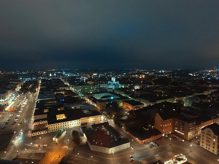 Ilmakuva ilta-aikaan Helsingin keskustasta. Rakennuksissa näkyy valoja, mutkittelevalla suurella tiellä kulkee joitain autoja.