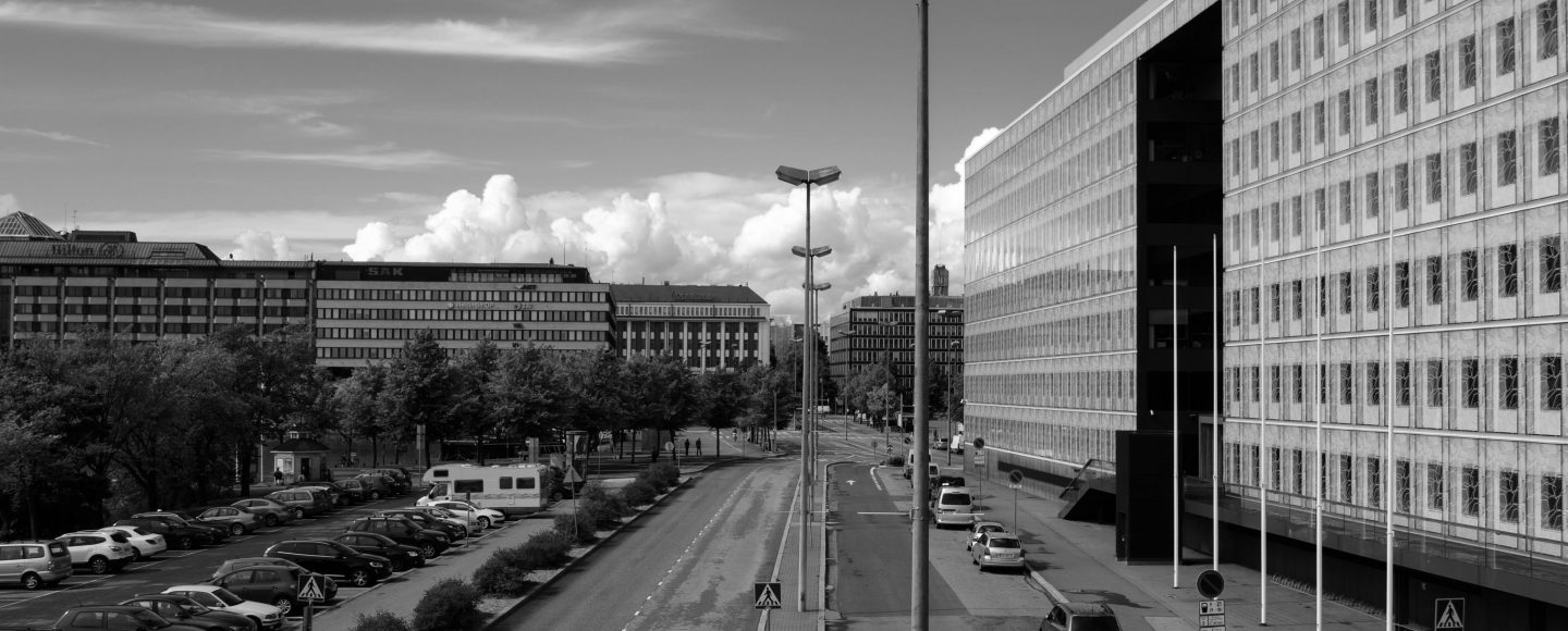 Mustavalkokuva kaupunkimaisemasta, jossa näkyy autotie, keskellä, kerrostalo oikealla ja puisto vasemmalla.