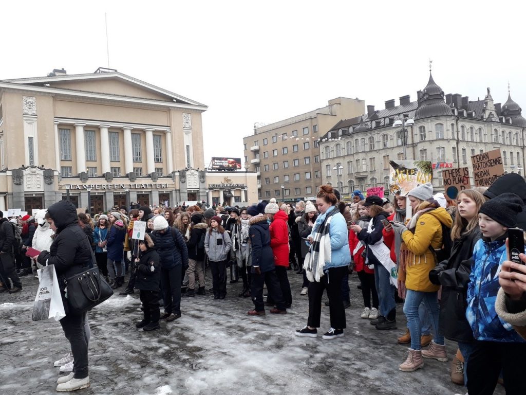 Mielenosoittajia seisoo talvivaatteissa kaupungissa. Takana on rakennuksia. Maassa on jäätä. Osalla mielenosoittajista on ympäristöaiheisia kylttejä käsissään.